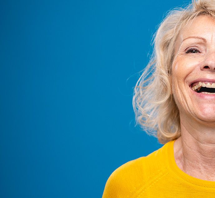 Female in her early 60s wearing a yellow jumper standing against a blue background